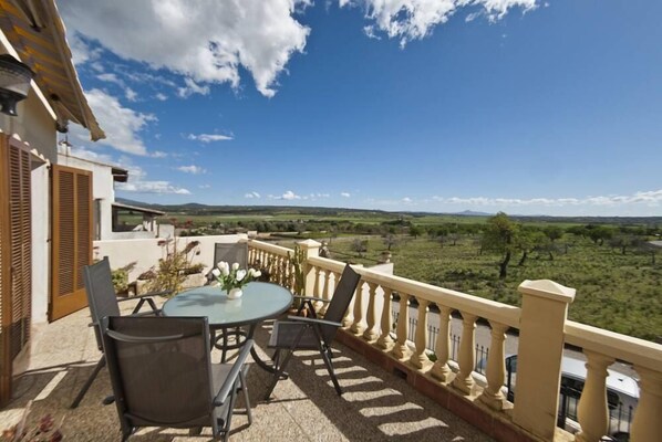 A balcony with a table and chairs, offering a view of a green landscape and distant hills.