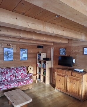 A cozy wooden cabin interior with a sofa, a small table, a TV, and a bookshelf.