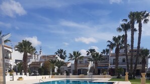 Una piscina con borde de baldosas, rodeada de palmeras y edificios con paredes blancas y techos de tejas rojas.