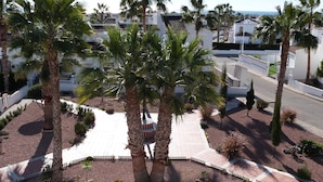 Zona ajardinada con palmeras, un sendero pavimentado y un edificio blanco con tejado de baldosas.