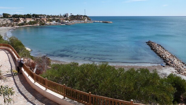 Vista costera con cielo despejado, un rompeolas rocoso y un paseo marítimo con barandilla.