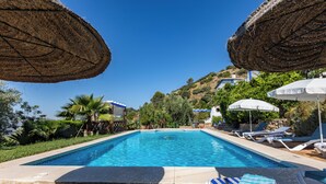 Une piscine entourée de verdure et d’une colline, avec des chaises longues et des parasols.