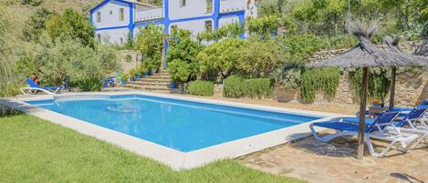A swimming pool with lounge chairs, a thatched-roof structure, and a house with blue and white exterior walls.