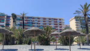 Ett område vid stranden med parasoller av halmtak, palmer och ett hotell med flera våningar och balkonger.