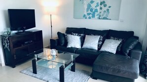 Una sala de estar moderna con una mesa de centro de vidrio, un sofá negro con cojines estampados, un televisor de pantalla plana sobre un soporte de madera y una obra de arte colgada en la pared.