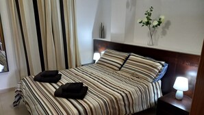 Una cama bien hecha con ropa de cama a rayas, una lámpara de noche, un jarrón con flores y un espejo en la pared.