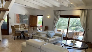 A spacious living area with a large sofa, dining table, and chairs. There is a balcony with a view of greenery. - Elegant Family Villa with Private Pool, Jacuzzi, Spacious Garden & Games Area (Crestatx -Sa Pobla)