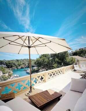Ein Balkon mit einem Holztisch, weißen Lounge-Stühlen und einem großen Sonnenschirm.
