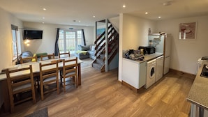 A modern kitchen with a built-in washing machine, a dining area with wooden furniture, and a staircase leading to another level. - Harlyn Bay Padstow, 70 Steps from the Beautiful Stunning Beach, Parking  Garden (harlyn bay padstow)
