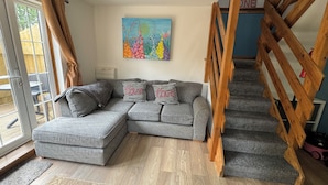 A modern living room with a grey sectional sofa, a colorful abstract painting, and a wooden staircase. - Harlyn Bay Padstow, 70 Steps from the Beautiful Stunning Beach, Parking  Garden (harlyn bay padstow)