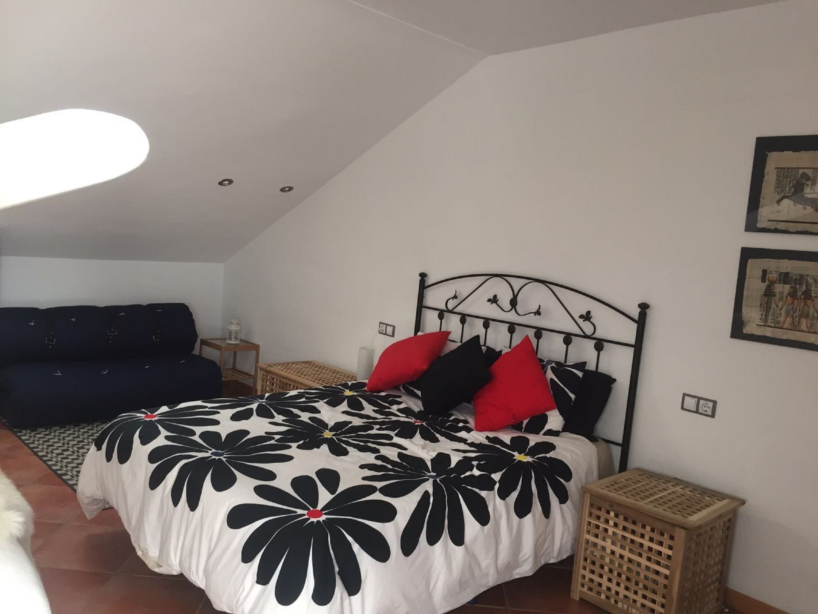 A bedroom with a bed, a nightstand, a lamp, a sofa, and a wall with pictures.
