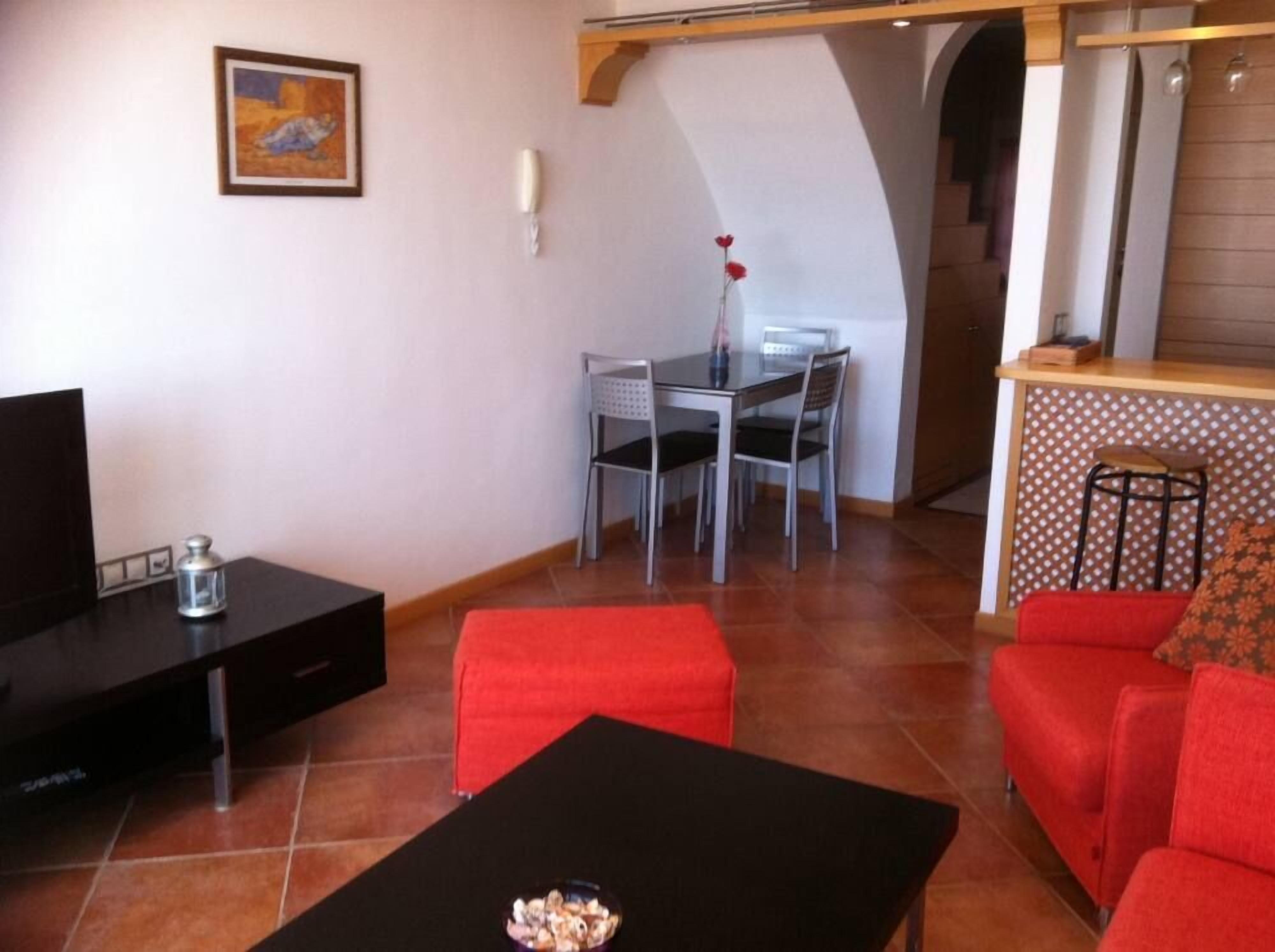 A living room with a TV, a dining table with chairs, a red sofa, and a kitchen area with a bar stool.