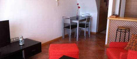A living room with a TV, a dining table with chairs, a red sofa, and a kitchen area with a bar stool.