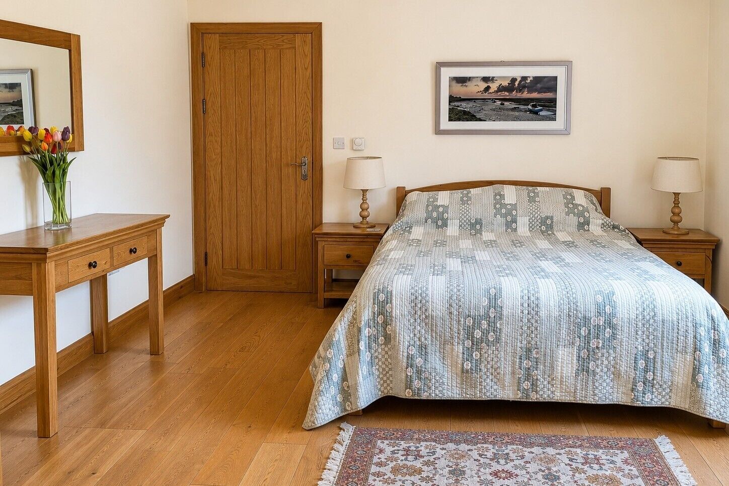 Een slaapkamer met een houten deur, een nachtkastje met een lamp, een bed met een beddengoed met dessin, een vloerkleed en een ingelijst kunstwerk aan de muur.