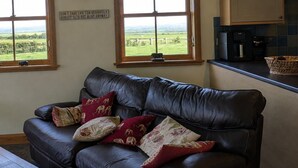 Ein Wohnzimmer mit einer Ledersofa, mehreren Kissen, einer Uhr und Blick auf eine grüne Landschaft durch große Fenster.