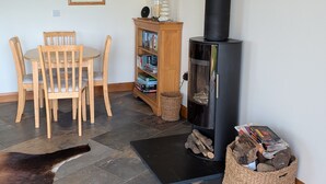 Ein gemütliches Zimmer mit einem runden Holztisch, vier Stühlen, einem Bücherregal, einem schwarzen Holzofen und einem gewebten Korb voller Holzscheite.