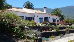 Um edifício branco com telhado em azulejos, rodeado por um jardim com diversas plantas e flores, e montanhas ao fundo.