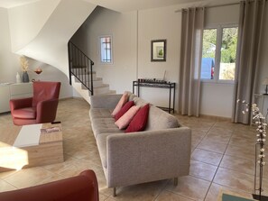 A living room with a sofa, a red chair, a staircase, a small table, and a window with curtains.