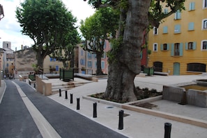 Une rue bordée d’arbres, avec des immeubles aux volets bleus et à la façade jaune.