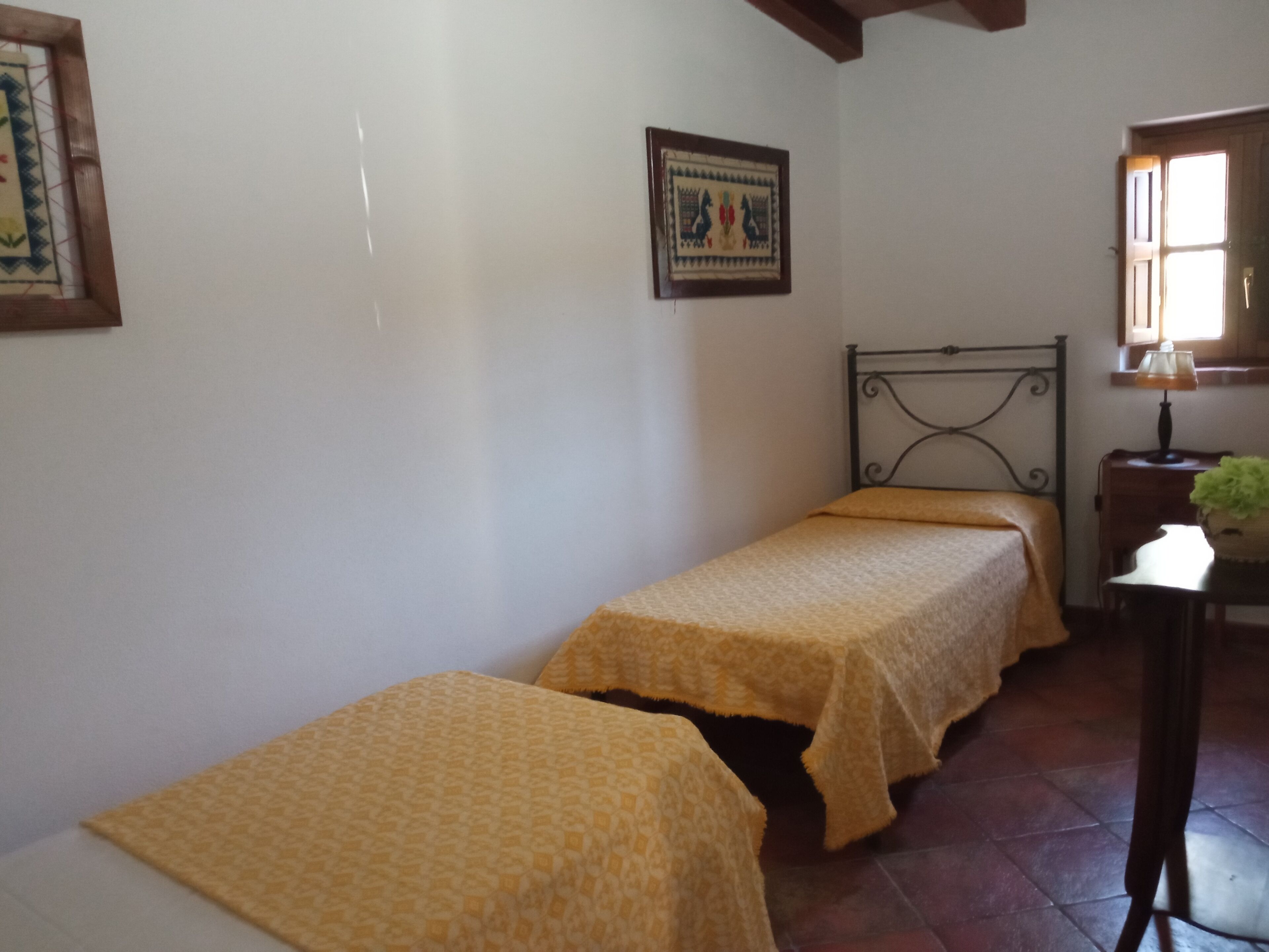 A small, simple room with two single beds, a wooden headboard, a small table with a lamp, and a window with natural light.
