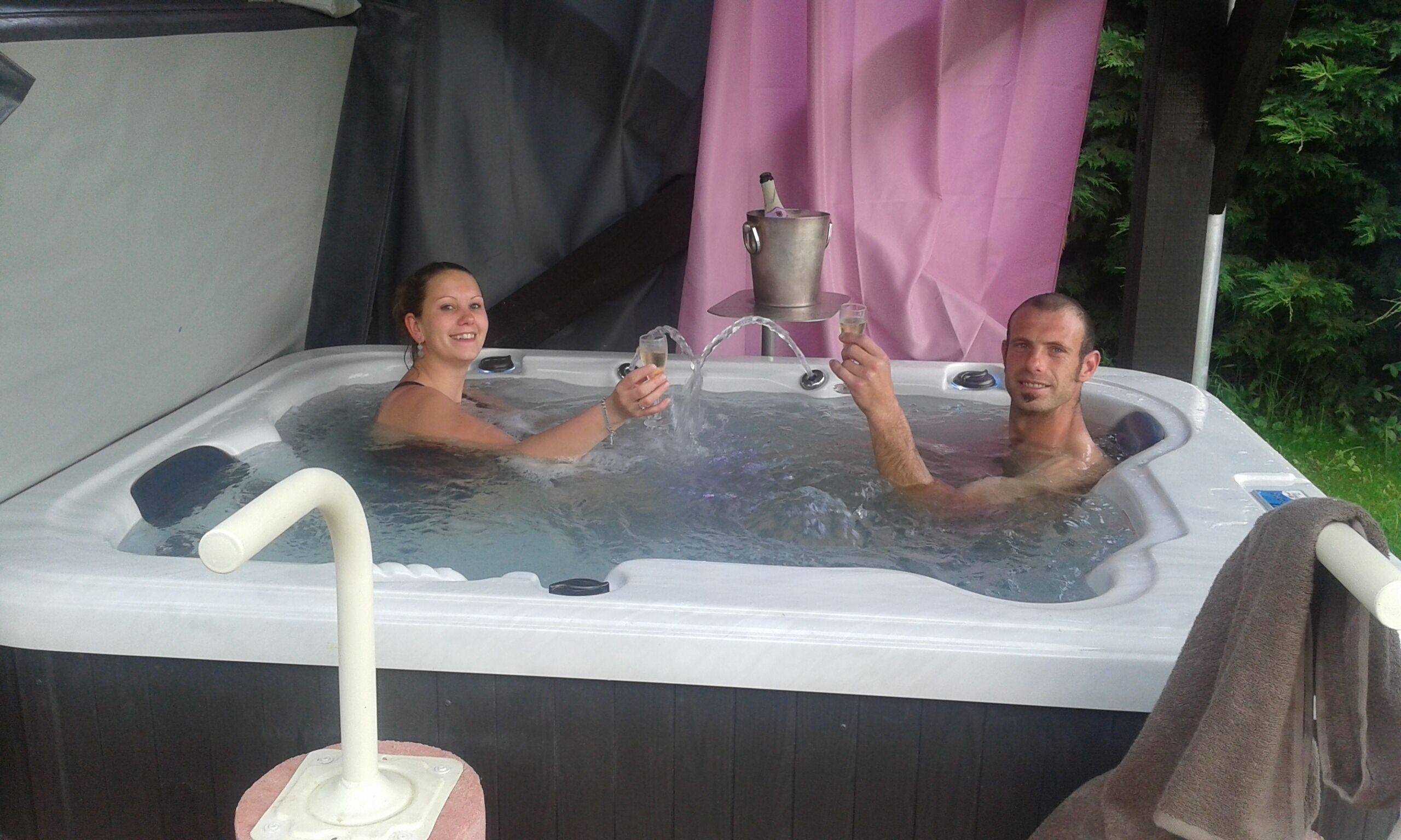 Een bubbelbad met twee personen die champagne glazen vasthouden, omringd door een roze gordijn en begroeiing.