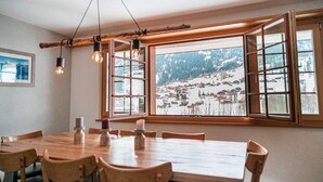 A dining area with a large wooden table, six chairs, and a view of a snowy mountain village through open windows. - Uninterrupted Eiger Views, 150m from Cable Car (Grindelwald)