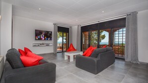 Un salon moderne avec des canapés gris et des coussins rouges, un téléviseur à écran plat et une grande fenêtre donnant sur la mer.