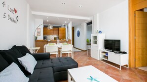 A modern living room with a black sofa, a white coffee table, and a TV on a white stand. There is a kitchen visible in the background with wooden cabinets and a dining area. - 3 bedroom apartment with sunny terrace perfect for families (Costa Adeje)