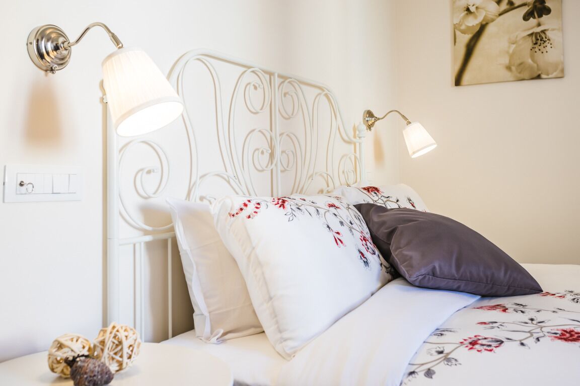 A neatly made bed with floral bedding, a white metal headboard, and two wall-mounted reading lamps.