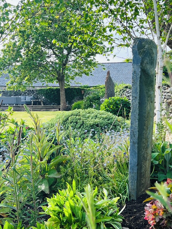 A garden with a stone pillar, various plants, and a building in the background. - Hen Stabl - A charming 18th century stone character stables conversion (Llanfair)