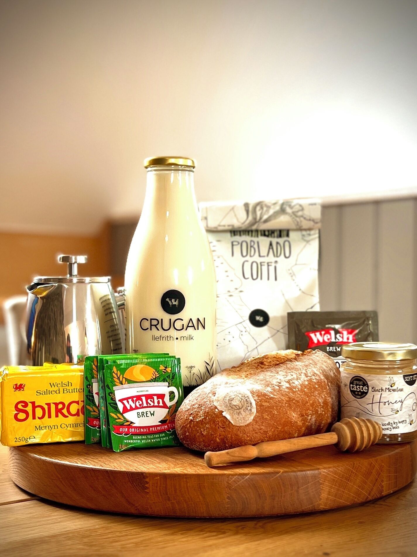 A wooden cutting board with a loaf of bread, a bottle of Crugan milk, Welsh Brew tea bags, a jar of honey, a packet of salted butter, and a box of Poblado Cotti coffee.