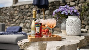 Una bandeja de madera con una botella de Aperol, una botella de vino tinto y dos vasos de un cóctel rosado con fresas sobre una mesa de piedra.