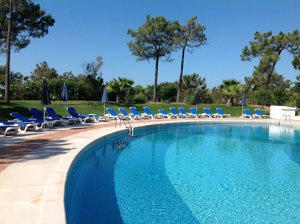 A pool area with blue lounge chairs and umbrellas, surrounded by trees and a clear sky.
