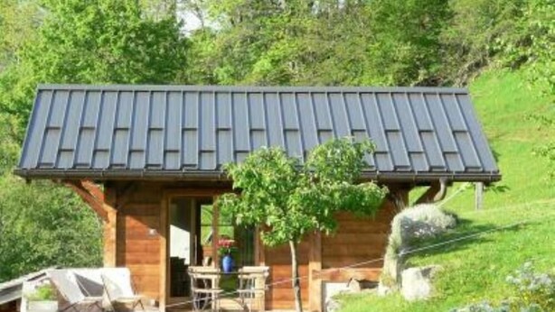 Une cabane en bois avec un toit en pente, une petite véranda et une table avec des chaises à l’extérieur.