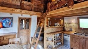 Un intérieur rustique en bois, avec un lit en mezzanine, une échelle, un coin cuisine doté d’étagères et une table à manger.