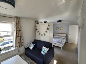 Una habitación pequeña con sofá, cama, ventana con cortinas, un adorno en la pared y una mesita.