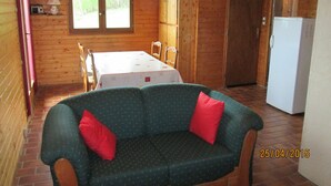 Een gezellig chalet met een eettafel, stoelen, een bank met rode kussens, een koelkast en een raam met uitzicht op bomen.