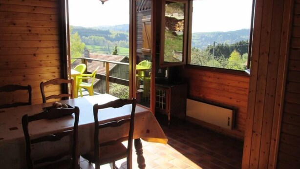 Een houten interieur met een eettafel, stoelen en een televisie. Grote ramen bieden uitzicht op een bergachtig landschap.