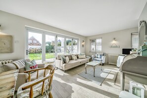 Ett rymligt vardagsrum med en soffa, ett soffbord och trästolar, stora fönster med utsikt över ett hus och en trädgård samt ett element.
