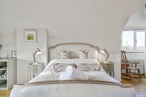 A bedroom with a large bed, two bedside lamps, a wooden rocking chair, a radiator, and a bookshelf.