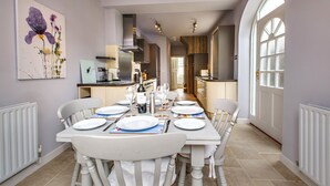 A dining area with a white table set for six, a kitchen in the background, and a large arched window on the right.