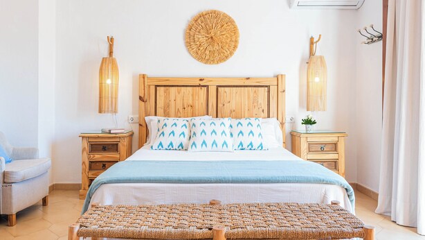 Un dormitorio con una cama de madera, dos mesitas de noche, un banco tejido, un adorno circular en la pared y una lámpara de pared.