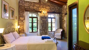 A bedroom with a stone wall, a bed with white bedding, two bedside tables with lamps, a chandelier, and a view through the window. - Blue Paradise villa With Pool And Jacuzzi For 5 People (rethymno)