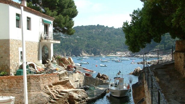 A coastal town with a narrow waterway, boats docked along the shore, and a stone building with a balcony.