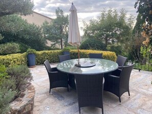 A round glass table with wicker chairs and a beige umbrella in a garden setting. - Hillside villa, magnificent views, heated private pool, short walk to village (Calonge)