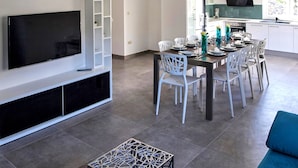 A modern living room with a flat-screen TV, white chairs, a dining table set for four, and a kitchen area with a built-in oven and microwave. - Lux villa,  heated infinity salt water pool, sauna, whirpool, close to the sea. (Smolonje)