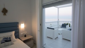 A bedroom with a bed, bedside table, and a view of a balcony with white wicker furniture and a railing.