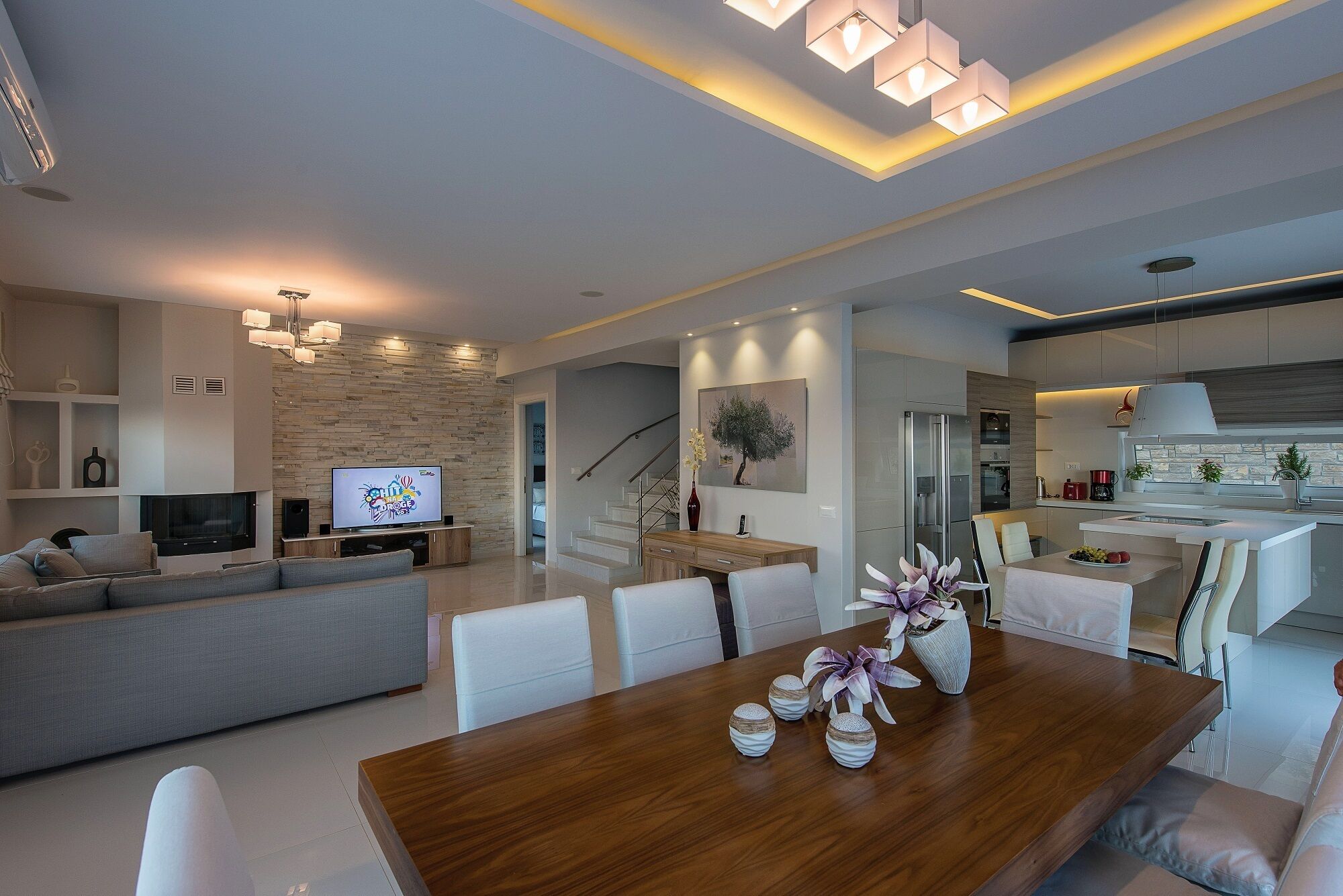 A modern living room with a wooden dining table, a sofa, and a TV. There is a staircase leading to another level.