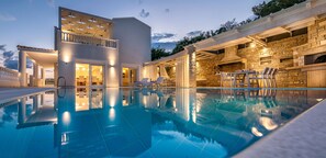 A modern poolside area with a clear pool, outdoor seating, and a stone fireplace. - Luxury villa,heating pool superior facilities ideal for families, free transfer (Achlia)