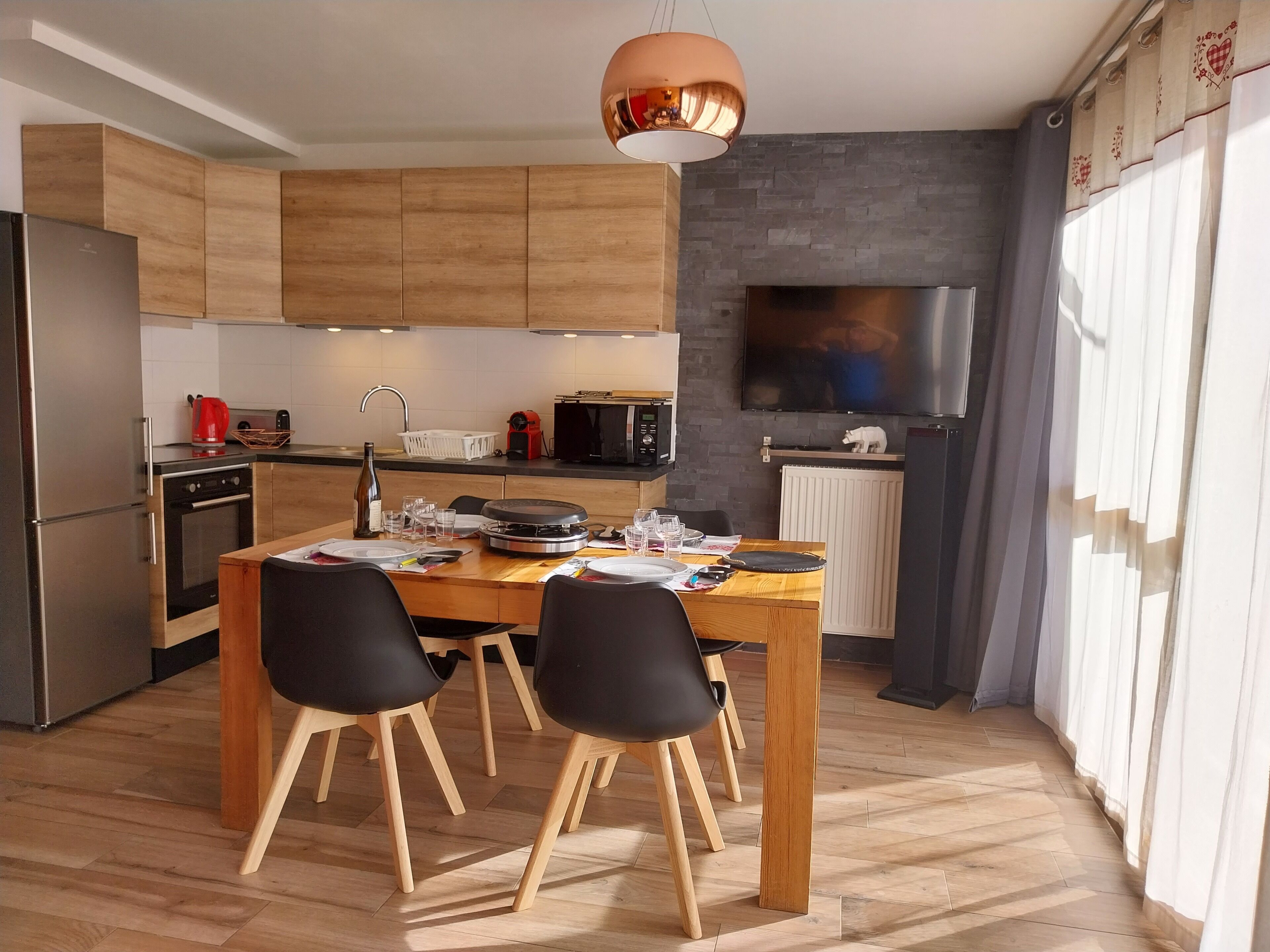 Une cuisine moderne avec des meubles en bois, une table à manger avec des chaises, un four encastré, un four à micro-ondes et un téléviseur fixé au mur.